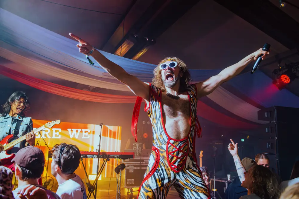 Man in tiger-striped outfit and white sunglasses sings energetically on stage holding a microphone.