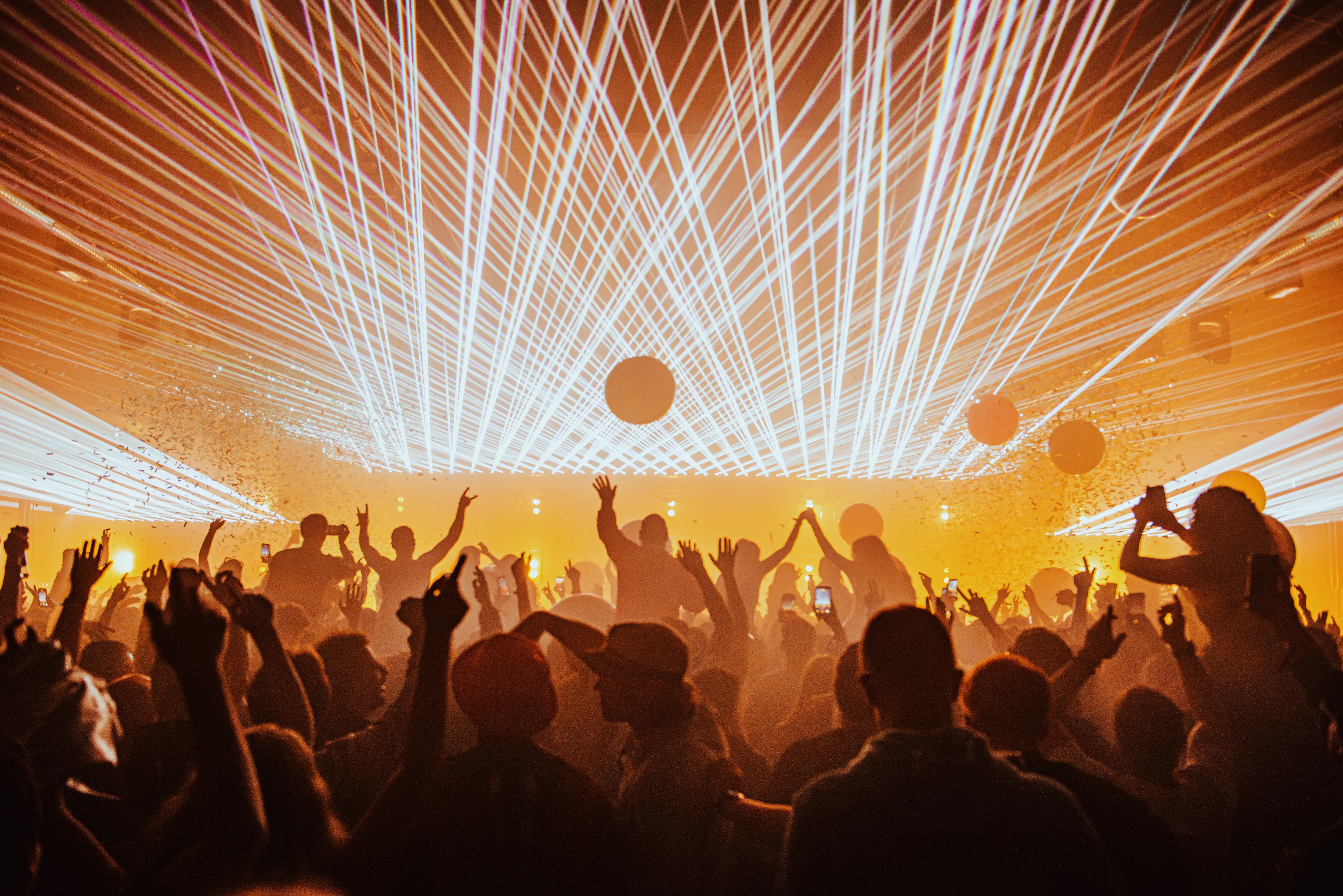 Crowd at a concert with raised hands, balloons, confetti, and bright white laser lights overhead.