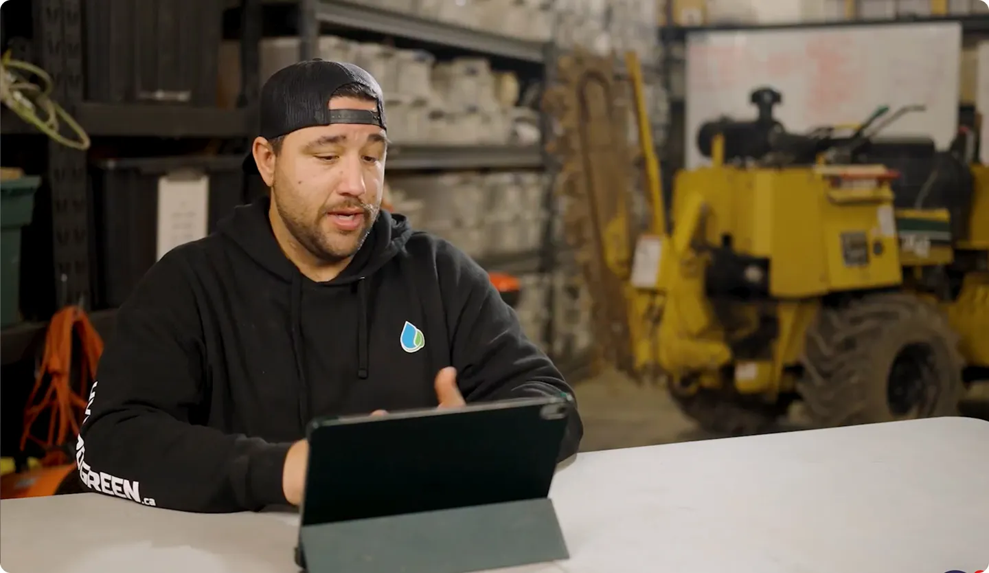 Man wearing a black hoodie and backwards cap sitting at a table using a tablet in an industrial workshop with shelves and machinery in the background.