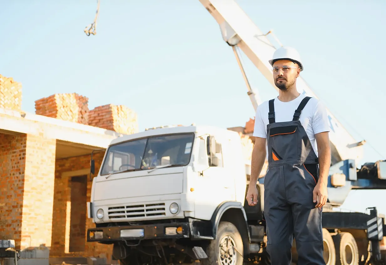 Construction worker wearing a white hard hat and safety glasses walking in front of a crane truck at a brick building construction site.