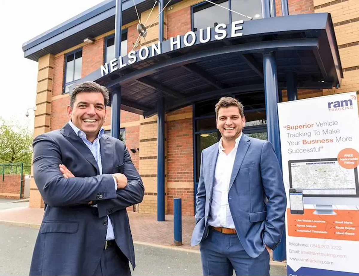 Two men in business suits smiling and standing outside the Nelson House building near a promotional sign for RAM Tracking.