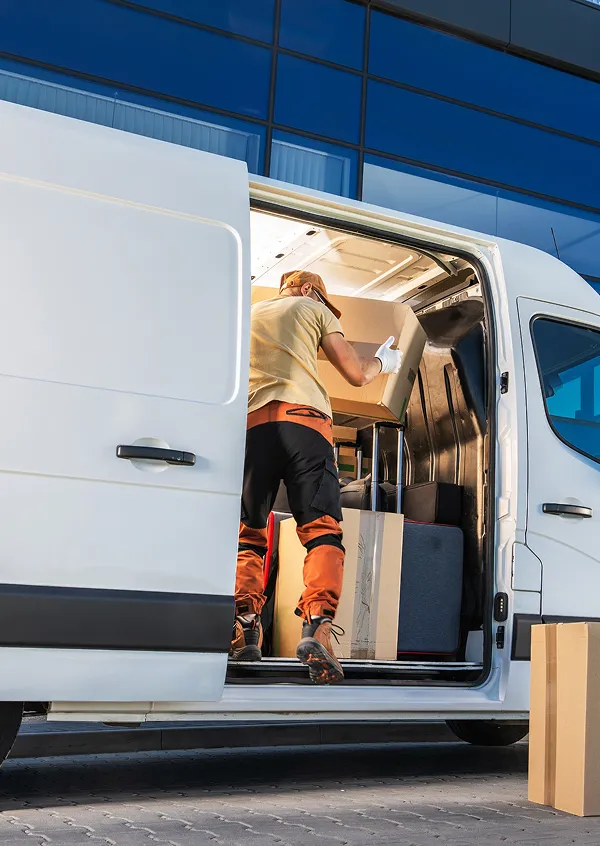 Delivery worker in orange and beige uniform loading cardboard boxes into a white van in front of a modern glass building.