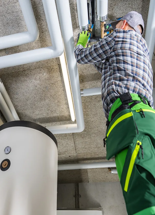 Technician in green pants and plaid shirt working on white insulated pipes near a large tank with gauge on a concrete ceiling.