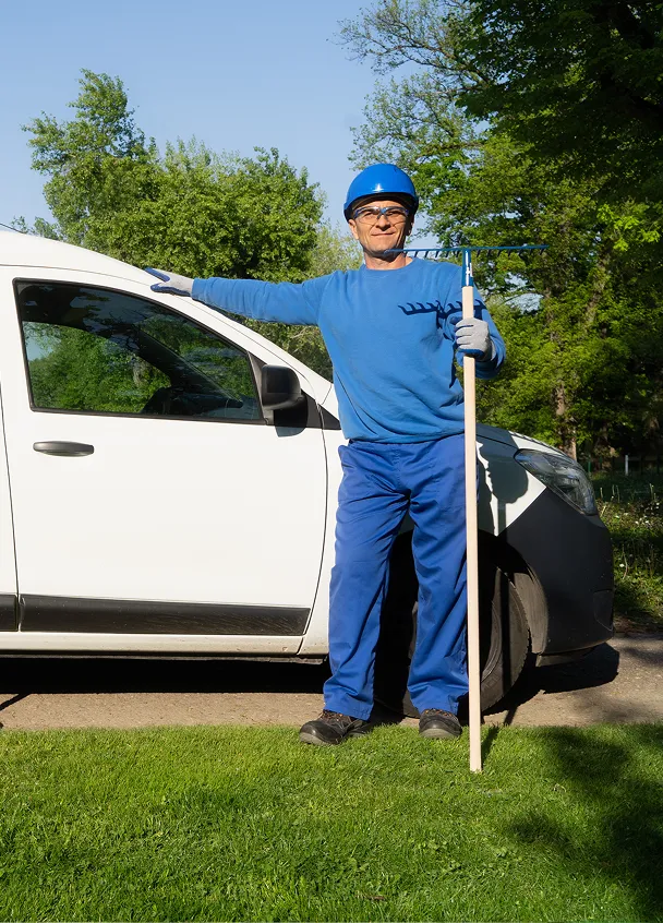 Worker in blue uniform and helmet holding a garden rake standing beside a white vehicle on grass.