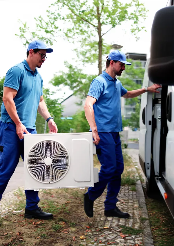 Two workers in blue uniforms carrying an air conditioning unit toward a white van outdoors.