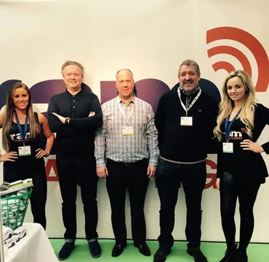Five people standing and posing indoors in front of a white wall with partial text and a red logo.