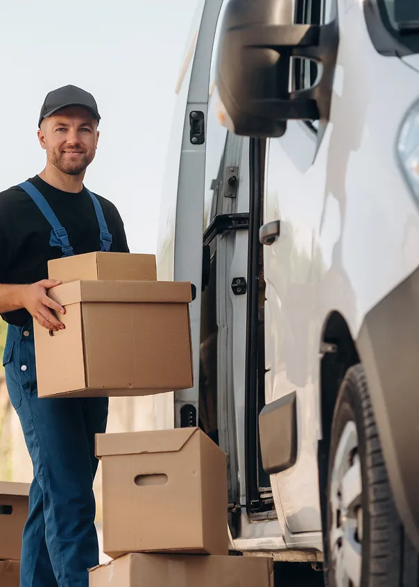 Delivery worker wearing blue overalls and black cap carrying cardboard boxes near a white van.