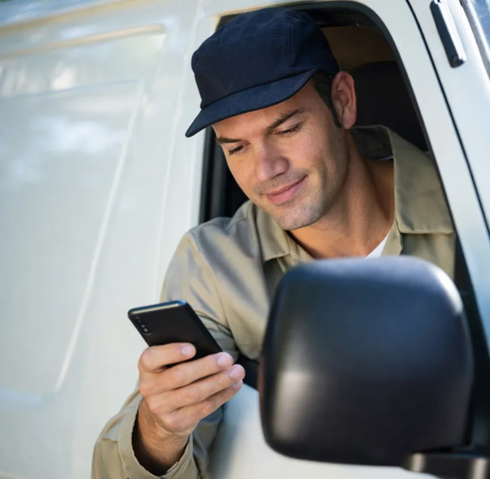 Man in a beige jacket and navy blue cap looking at a smartphone while sitting in the driver's seat of a white van.