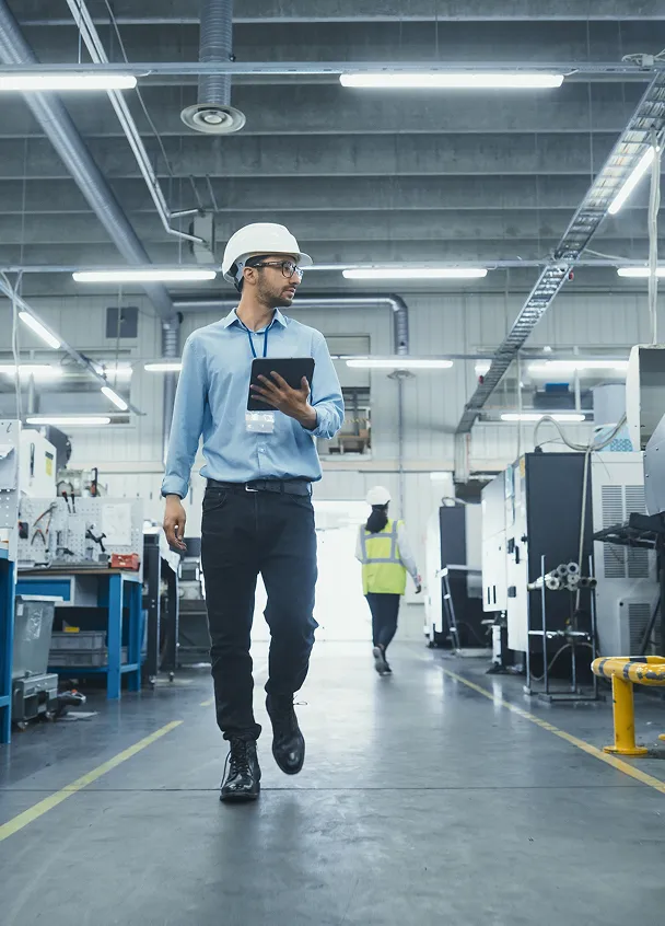 Man wearing a white hard hat and blue shirt walking in a factory while holding a tablet.