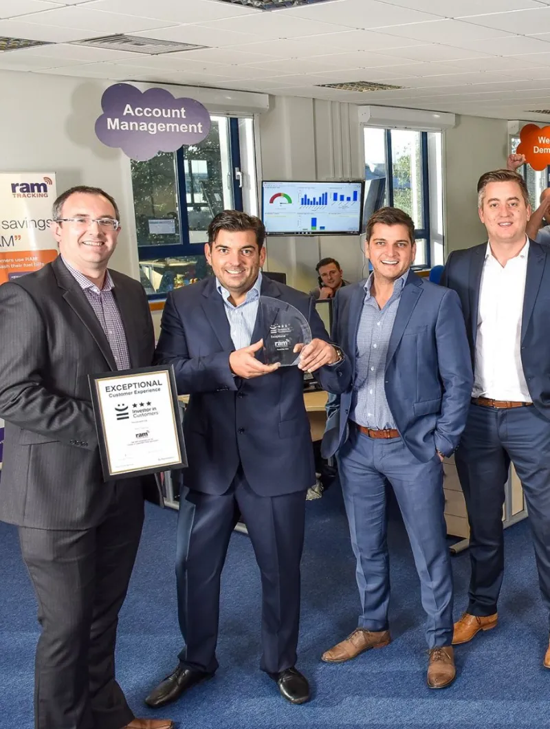 Four men in business attire standing in an office, two holding awards for exceptional customer experience from ram tracking.