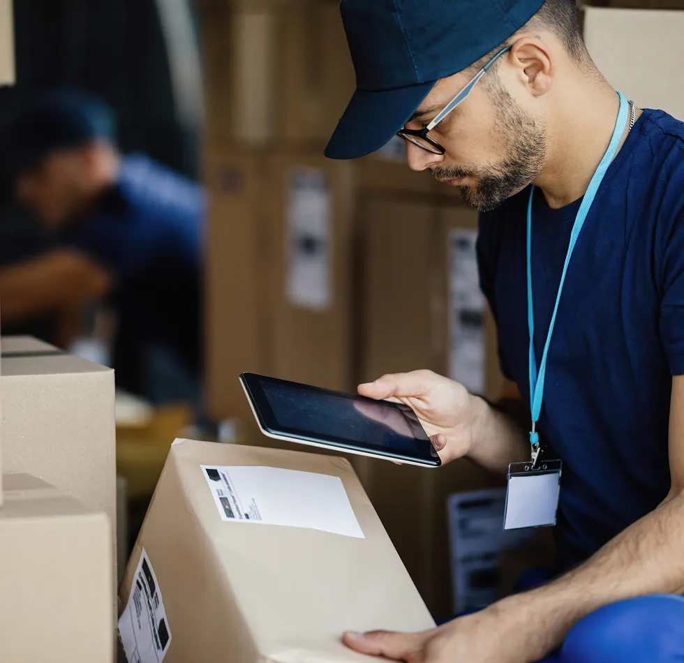 Man in a navy cap and shirt holding a tablet while inspecting a labeled cardboard box in a warehouse.