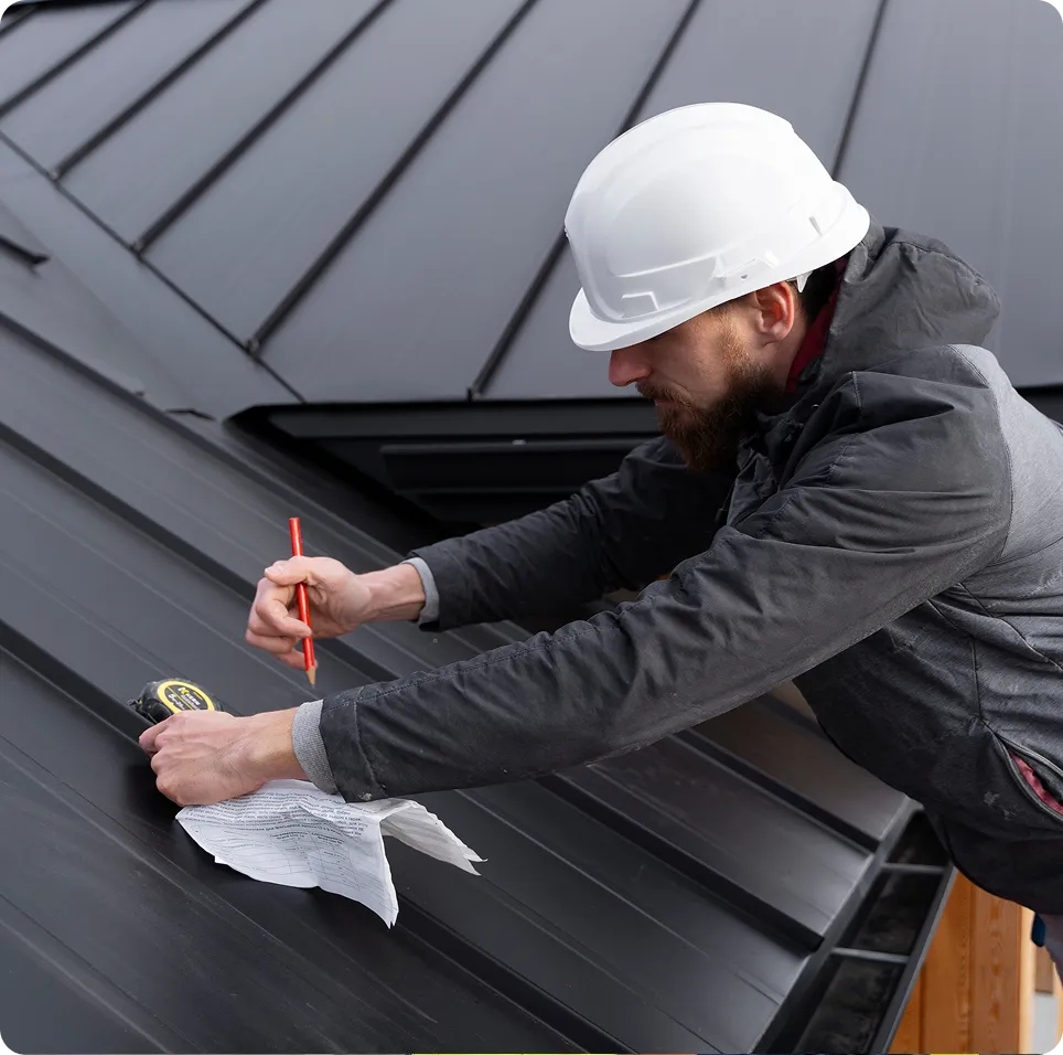 Construction worker wearing a white hard hat measuring and marking on a black metal roof using a tape measure and pencil.
