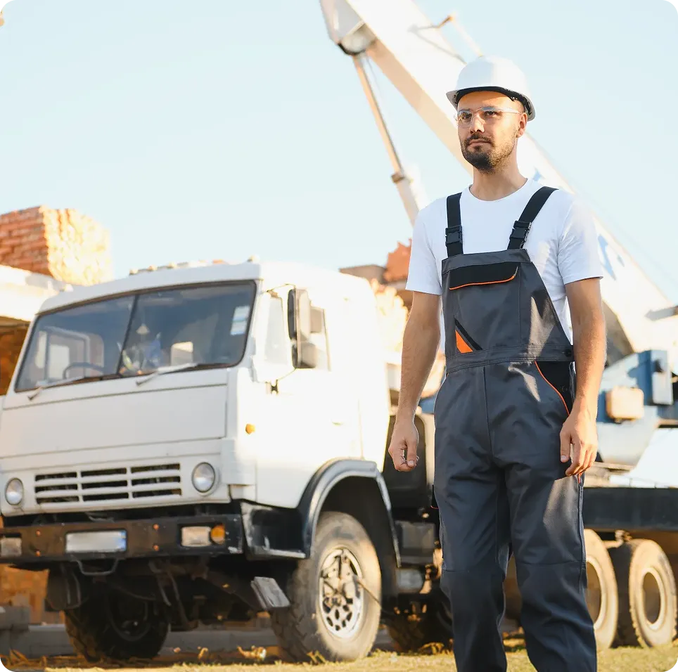 Construction worker wearing a white helmet and gray overalls standing near a white construction truck at a building site.