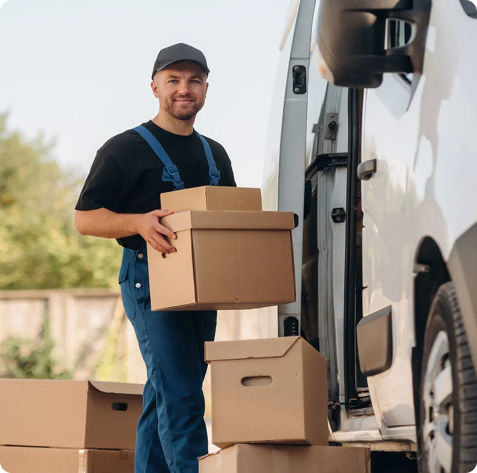 Delivery worker wearing blue overalls and black cap holding cardboard boxes near a white delivery van.