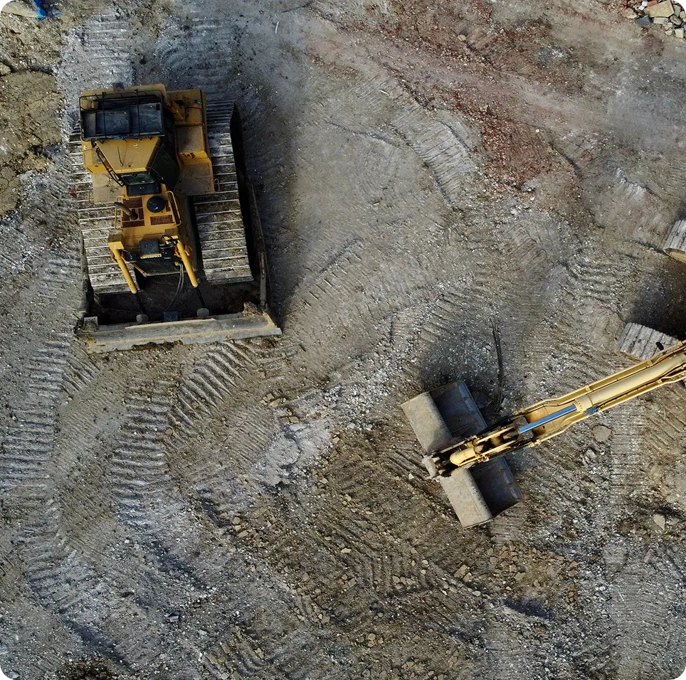 Aerial view of a construction site showing a yellow bulldozer and an excavator on dirt ground with tire tracks.