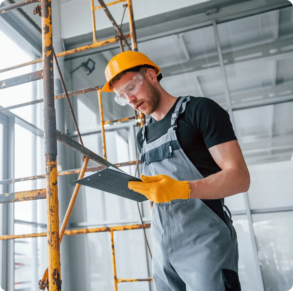 Construction worker wearing a yellow hard hat and protective glasses checking a clipboard near scaffolding indoors.