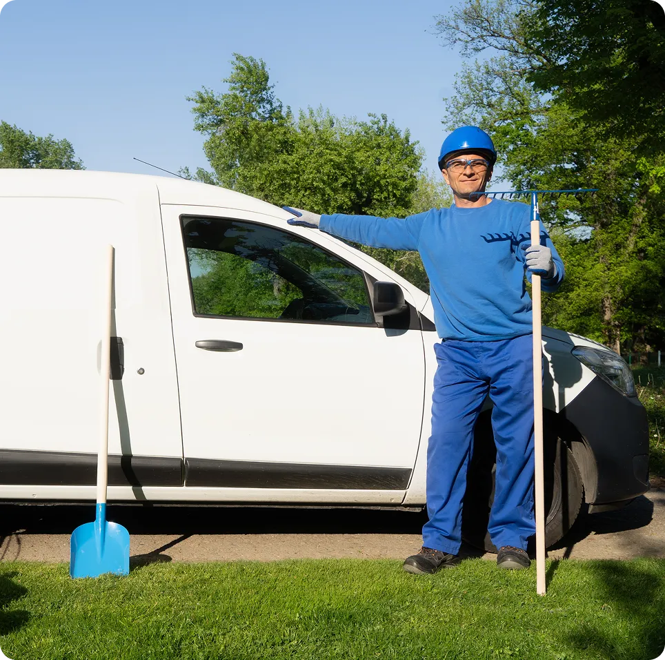 Gardener in blue workwear and helmet standing beside a white van holding a rake with a blue shovel leaning nearby on grass.