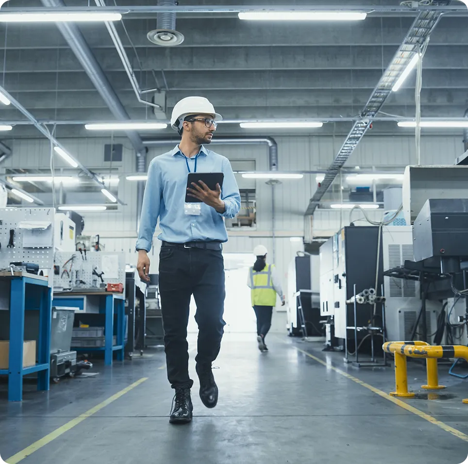 Man in a blue shirt and white hard hat holding a tablet walking in a modern industrial factory.