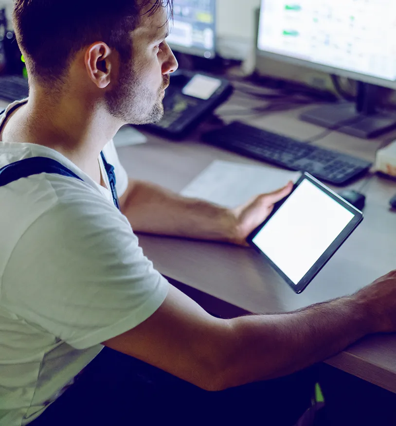 Man in white shirt holding a tablet with a blank screen, seated at a desk with computer monitors and keyboard.