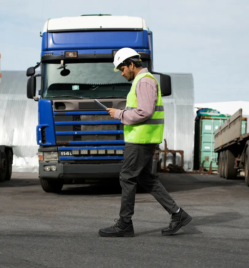 Worker in a safety vest and hard hat walking in front of a blue large truck while checking a clipboard.