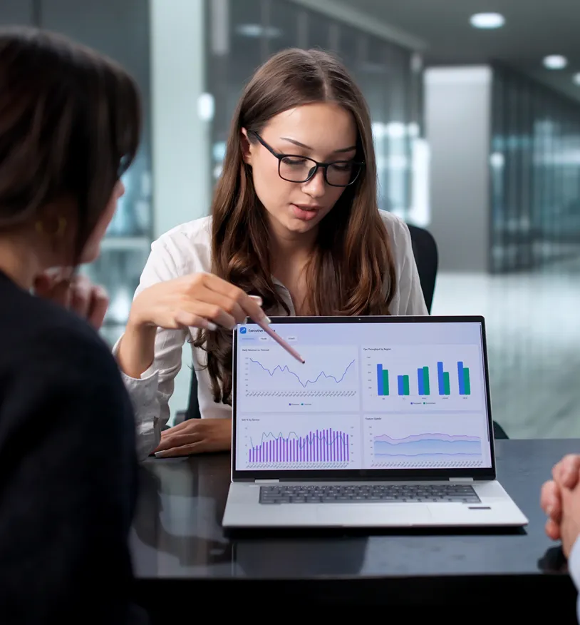Woman wearing glasses pointing at graphs and charts displayed on a laptop during a meeting with colleagues.