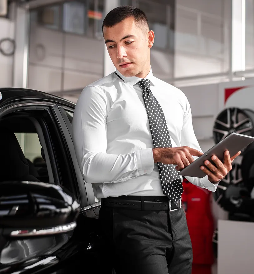 Man in white shirt and polka dot tie using a tablet while leaning on a black car in a showroom.