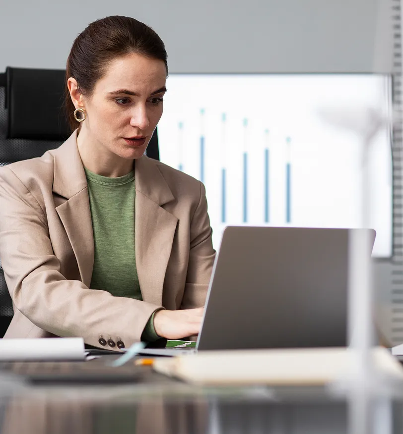 Woman in a beige blazer and green shirt working attentively on a laptop in an office with a bar chart displayed on a screen behind her.