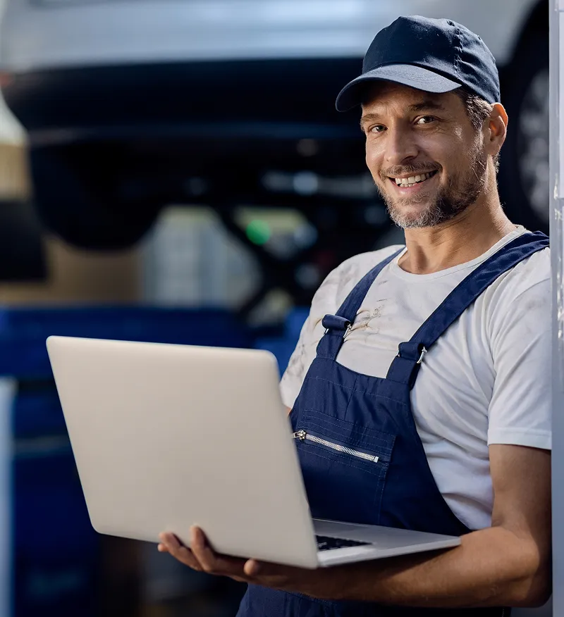 Smiling mechanic in overalls holding a laptop in a garage or workshop.