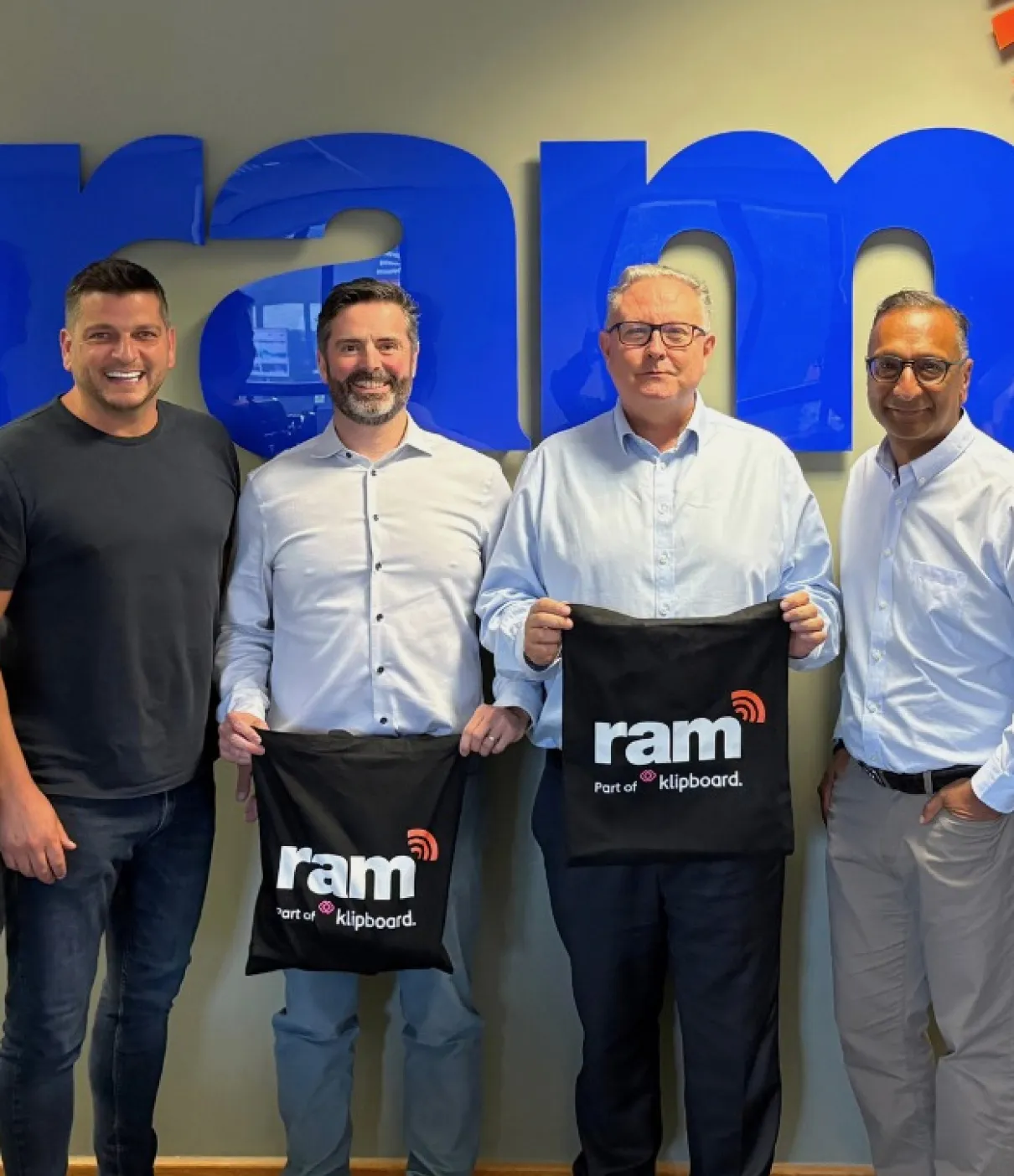 Four men standing in front of a large blue 'ram' sign, two holding black bags with 'ram part of klipboard' logos.