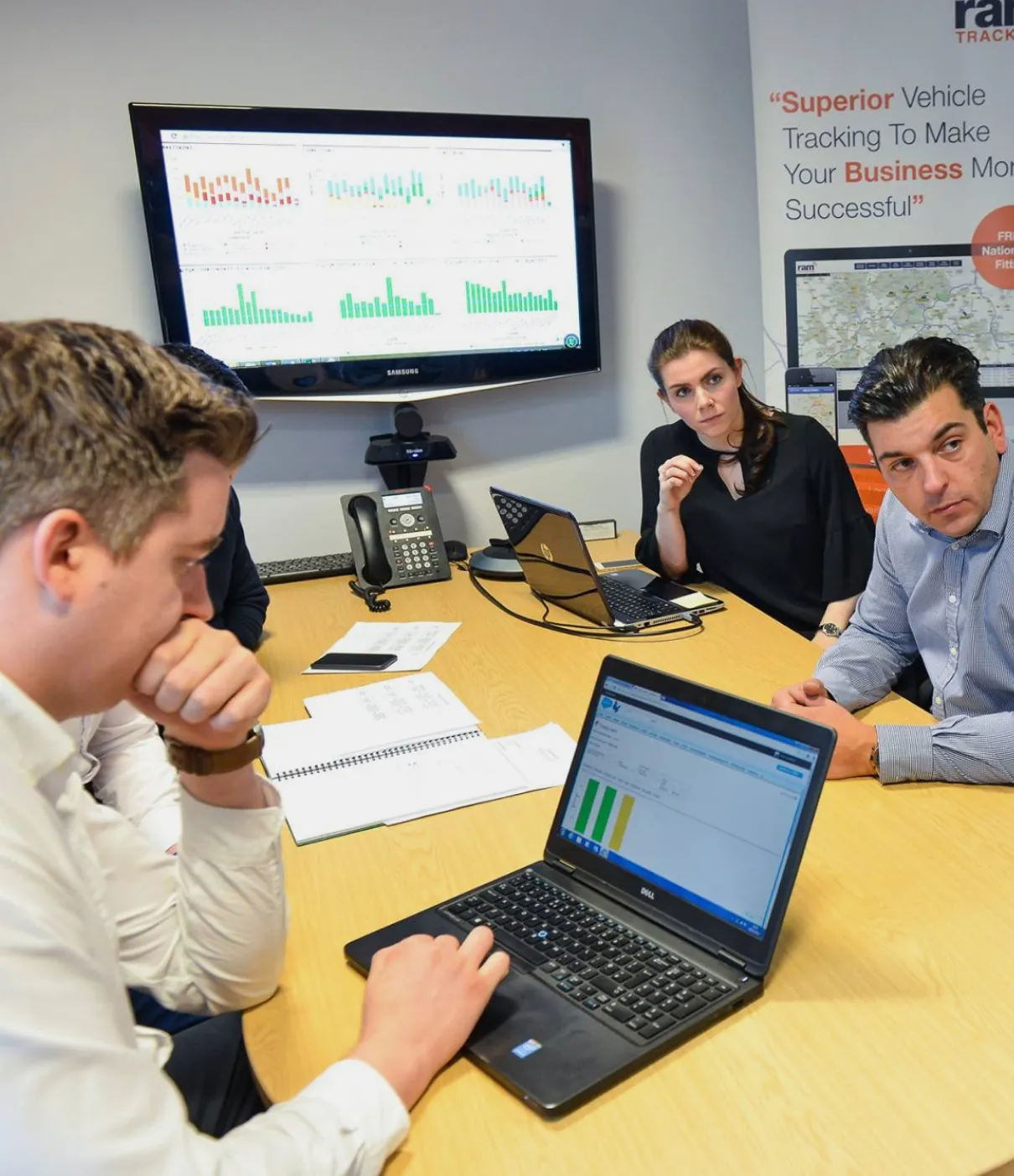 Four coworkers in a meeting room with laptops and charts displayed on a wall monitor and table discussing data insights.