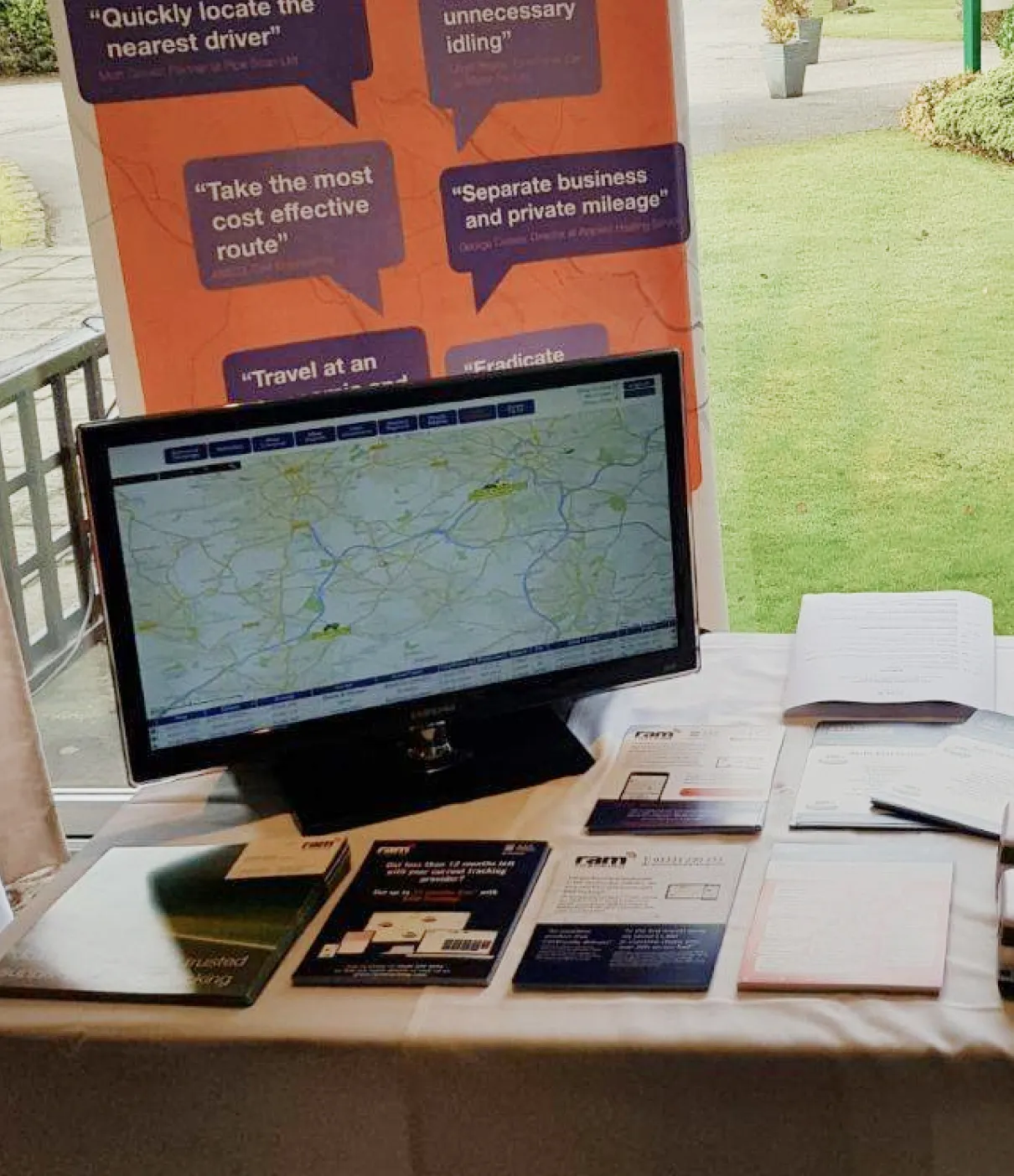 Table display with a computer monitor showing a map and various brochures and documents, with an orange poster in the background listing business mileage and driver management tips.