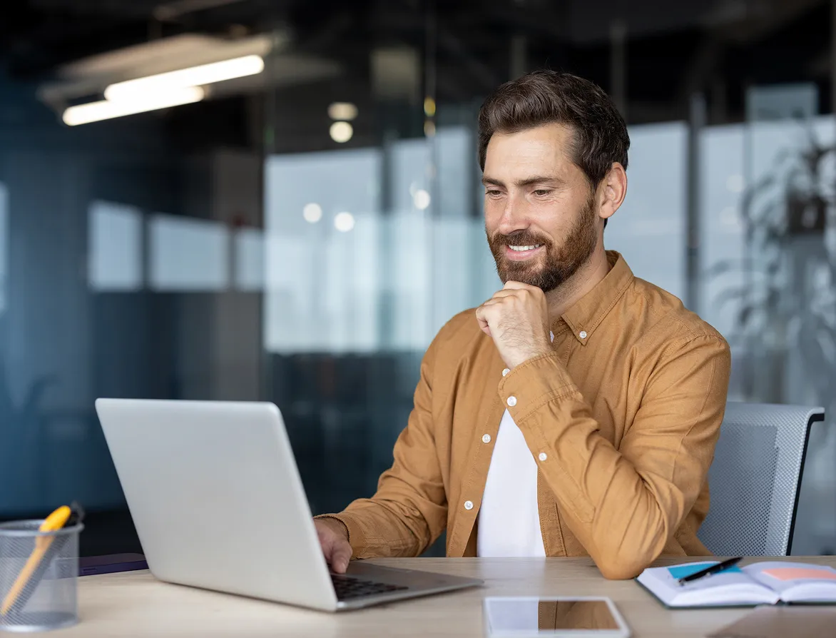 Smiling man with a beard in a brown shirt working on a laptop at a desk in a modern office.