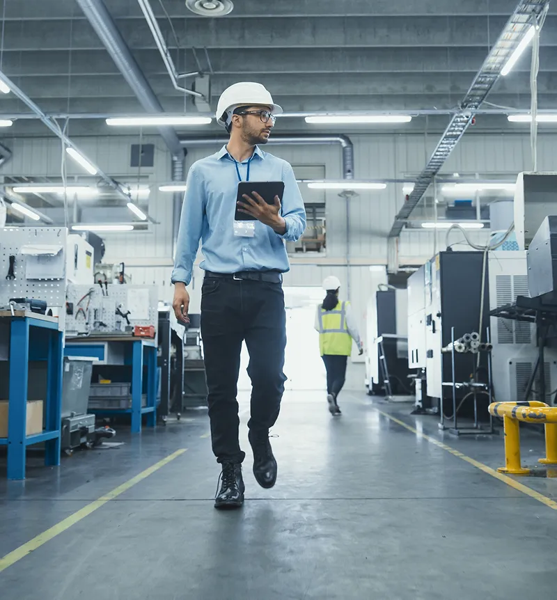Engineer wearing a white hard hat and glasses walking inside a factory while holding a tablet.