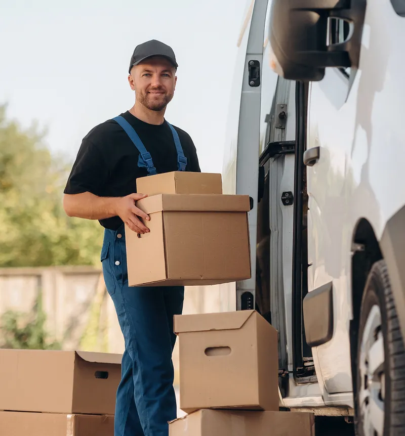 Delivery worker in blue overalls and black cap holding cardboard boxes next to a white van.