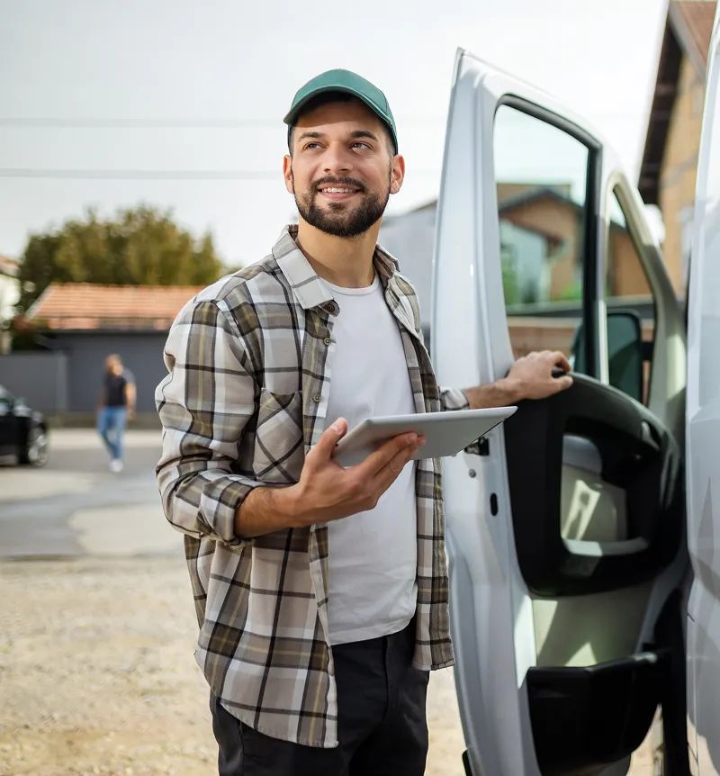 Man in plaid shirt and green cap holding a tablet standing next to an open van door outdoors.