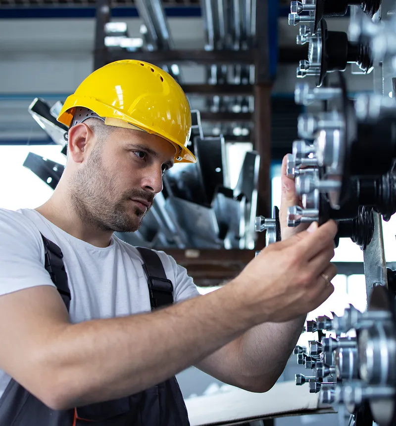 Worker wearing a yellow safety helmet adjusting industrial machinery bolts in a factory setting.