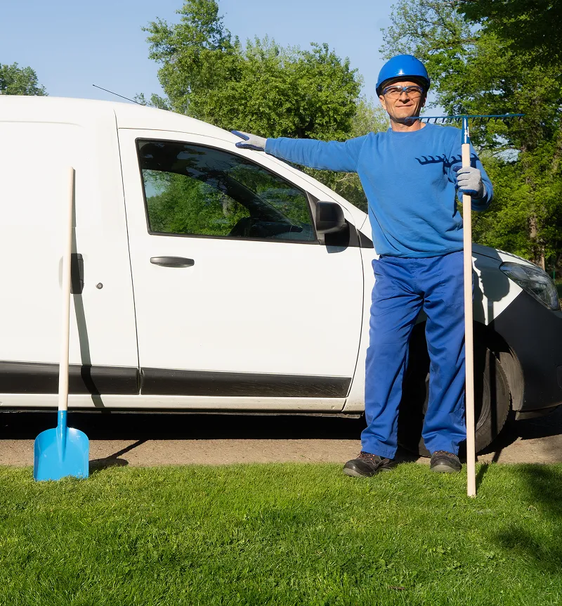 Gardener in blue workwear and helmet standing beside a white van holding a rake, with a blue shovel planted in the grass nearby.