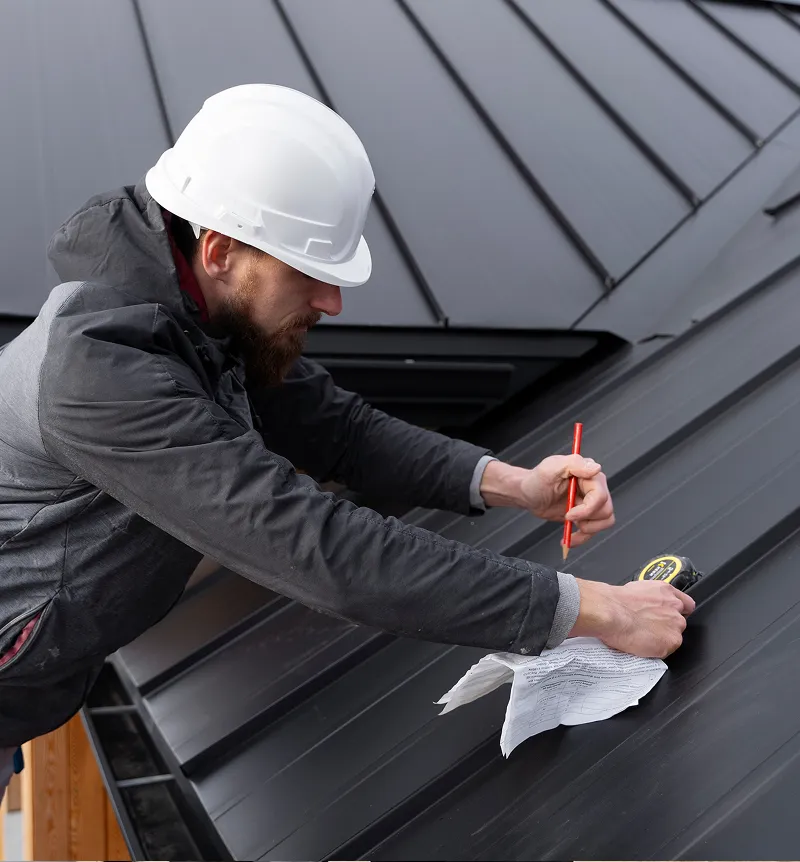 Construction worker wearing a white hard hat measuring and marking a dark metal roof panel with a tape measure and pencil.