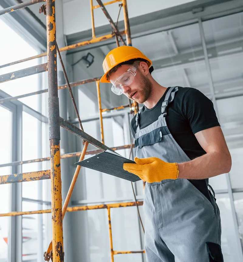 Construction worker wearing a yellow hard hat and safety goggles reading a clipboard near scaffolding inside a building.