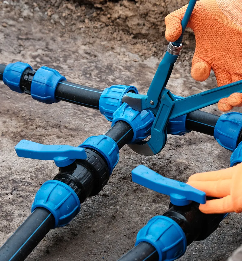 Hands wearing orange gloves using a blue pipe cutter to cut black irrigation pipes with blue fittings on soil.