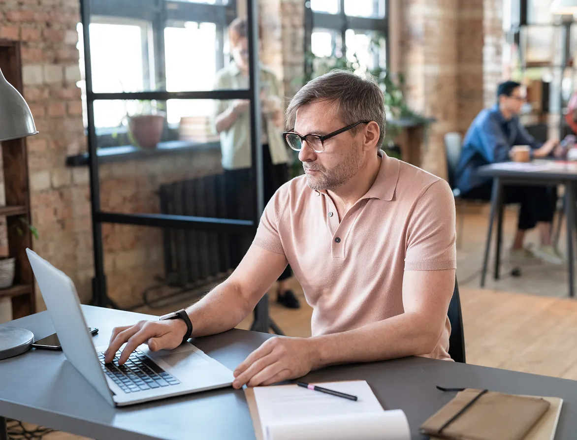 Man with glasses working on a laptop at a desk in a modern office with people and large windows in the background.