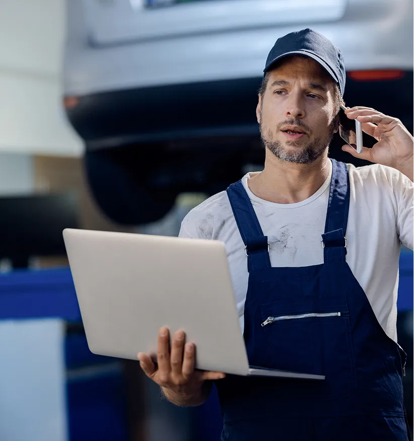 Mechanic on the phone holding a laptop in a vehicle workshop
