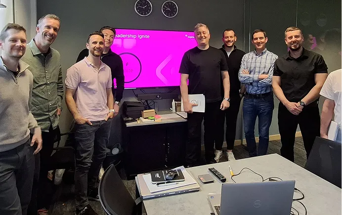 Eight men standing in an office meeting room with a screen showing 'Leadership Ignite' in the background.