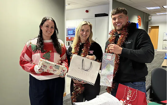 Three smiling people with tinsel hold wrapped Christmas gifts indoors.