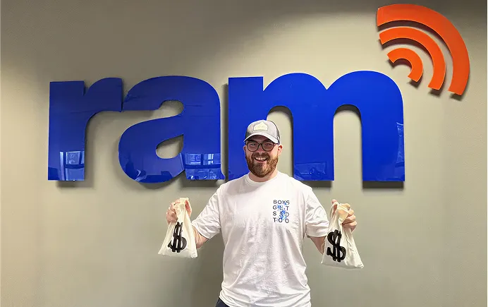 Man wearing glasses and caped smiling holding two white bags with black dollar signs in front of blue 'ram' sign.