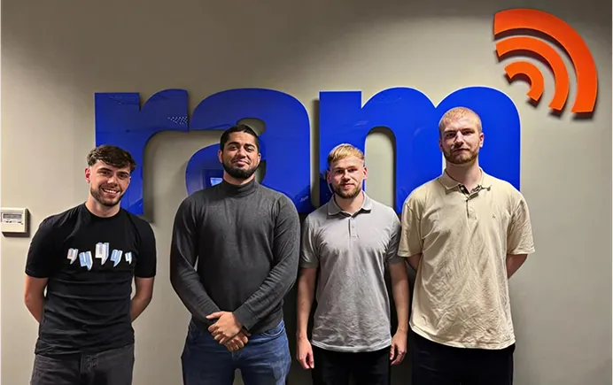 Four men standing smiling in front of a wall with a large blue and orange 'ram' logo.