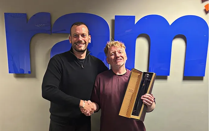 Two men shaking hands in front of blue letters, one holding a boxed award or gift.