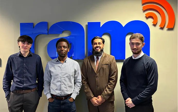 Four men standing in front of a wall with large blue letters spelling 'ram' and red signal waves above.