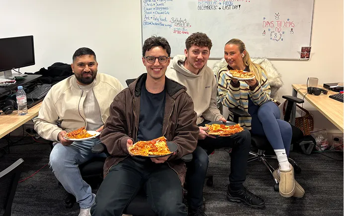 Four young people sitting in office chairs eating pizza, smiling at the camera with a whiteboard behind them.
