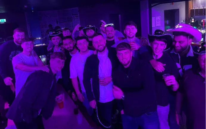 Group of young men posing together in a dimly lit bar under purple lighting, some wearing hats and holding drinks.